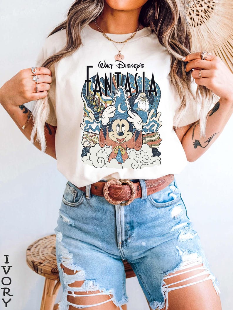 May include: Ivory t-shirt with a vintage graphic of Mickey Mouse from Disney's Fantasia. The design includes the text "Walt Disney's Fantasia" in a stylized font, with Mickey wearing a sorcerer's hat. The shirt is worn with denim shorts.