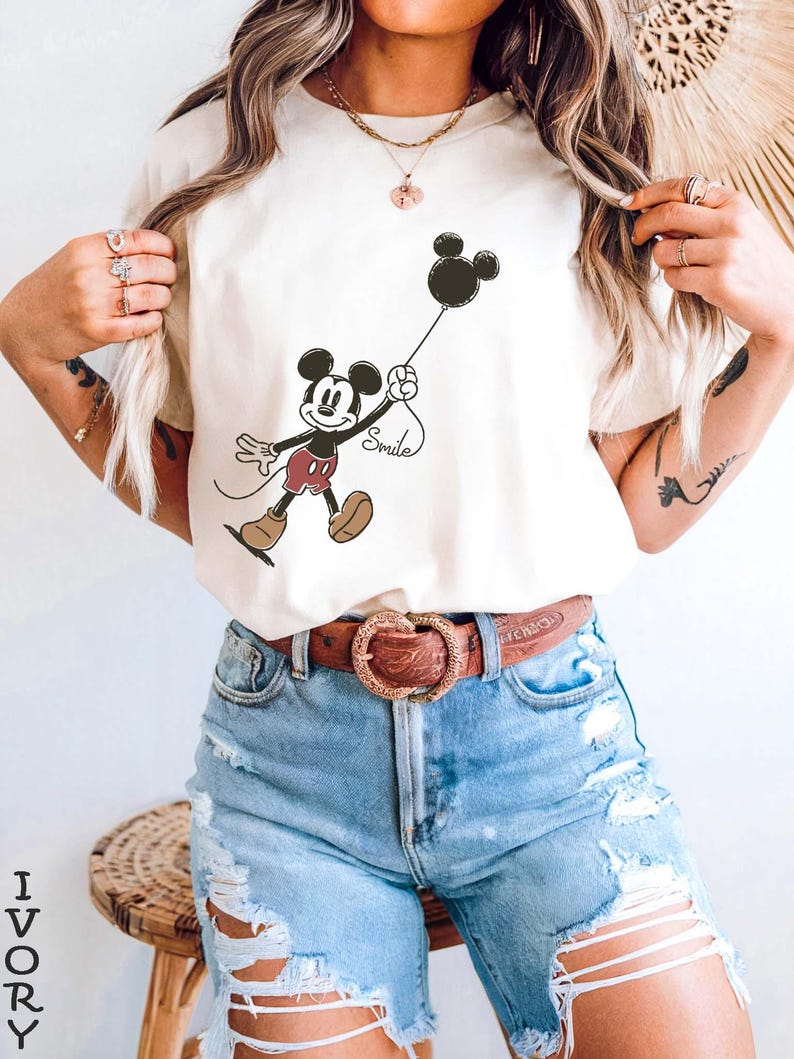 May include: Ivory t-shirt with a Mickey Mouse graphic holding a black balloon with "Smile" on the string. Mickey wears red shorts and brown shoes. The shirt is worn with light blue denim shorts. The word "IVORY" is printed vertically on the left.