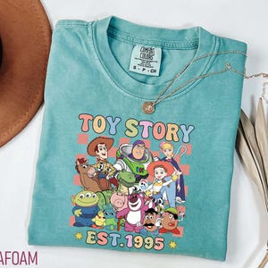 Może przedstawiać: Turkusowa koszulka z nadrukiem postaci z Toy Story, w tym Woody'ego, Buzza Astrala i Jessie. Nadruk otoczony jest r&oacute;żowo-ż&oacute;łto-niebieskim wzorem w kratkę. Pod nadrukiem znajduje się napis "TOY STORY EST. 1995".