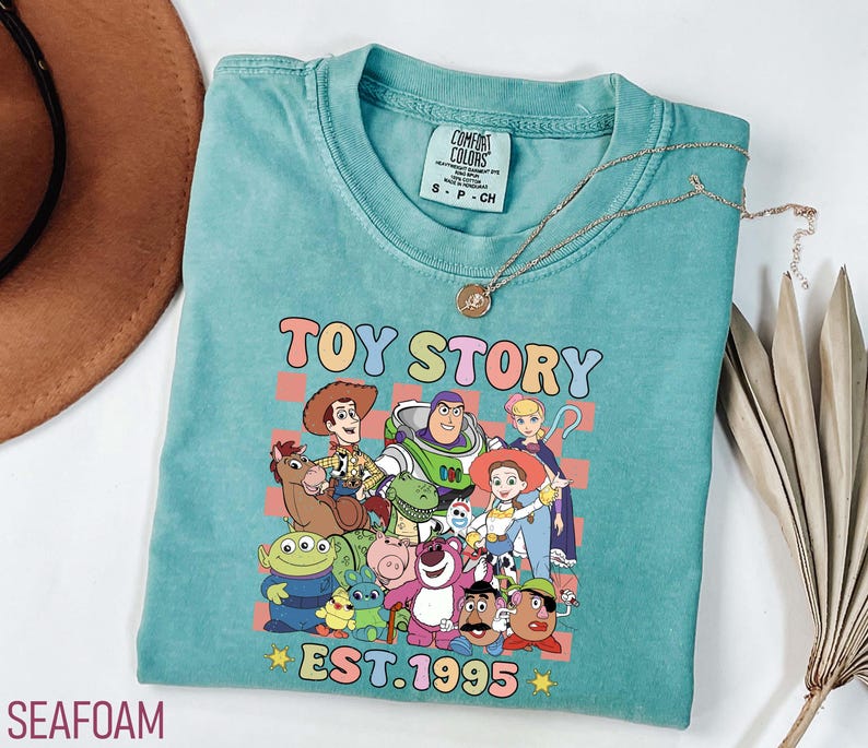 Może przedstawiać: Turkusowa koszulka z nadrukiem postaci z Toy Story, w tym Woody'ego, Buzza Astrala i Jessie. Nadruk otoczony jest r&oacute;żowo-ż&oacute;łto-niebieskim wzorem w kratkę. Pod nadrukiem znajduje się napis "TOY STORY EST. 1995".