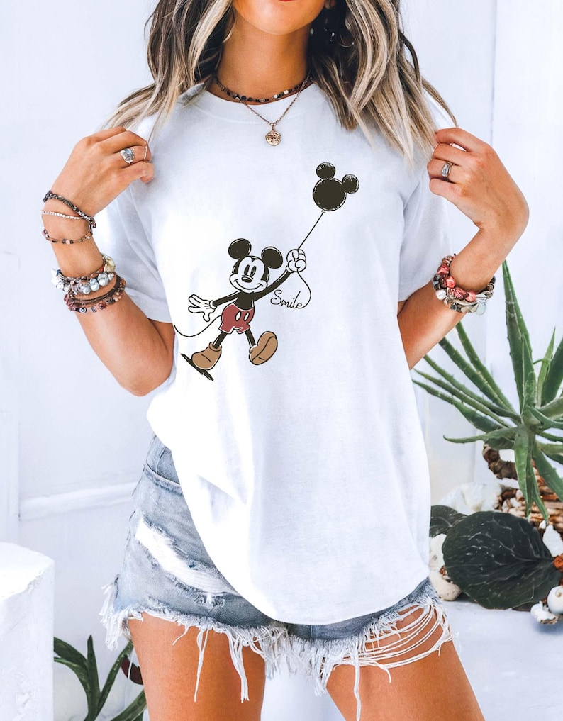 May include: White t-shirt featuring a cartoon Mickey Mouse holding a black balloon with the word "Smile" written below. Mickey is wearing red shorts and brown shoes. The shirt is a casual style.