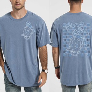 May include: Blue jean-colored t-shirt featuring a Donald Duck graphic on the front and back. The front graphic is a detailed outline of Donald Duck. The back graphic includes the words "Donald Duck" and a checkered pattern.