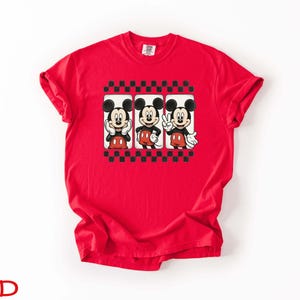 May include: A red t-shirt featuring three Mickey Mouse images in a black and white checkered frame. Mickey is depicted in various poses, wearing his signature red shorts and white gloves. The word "RED" is printed at the bottom.