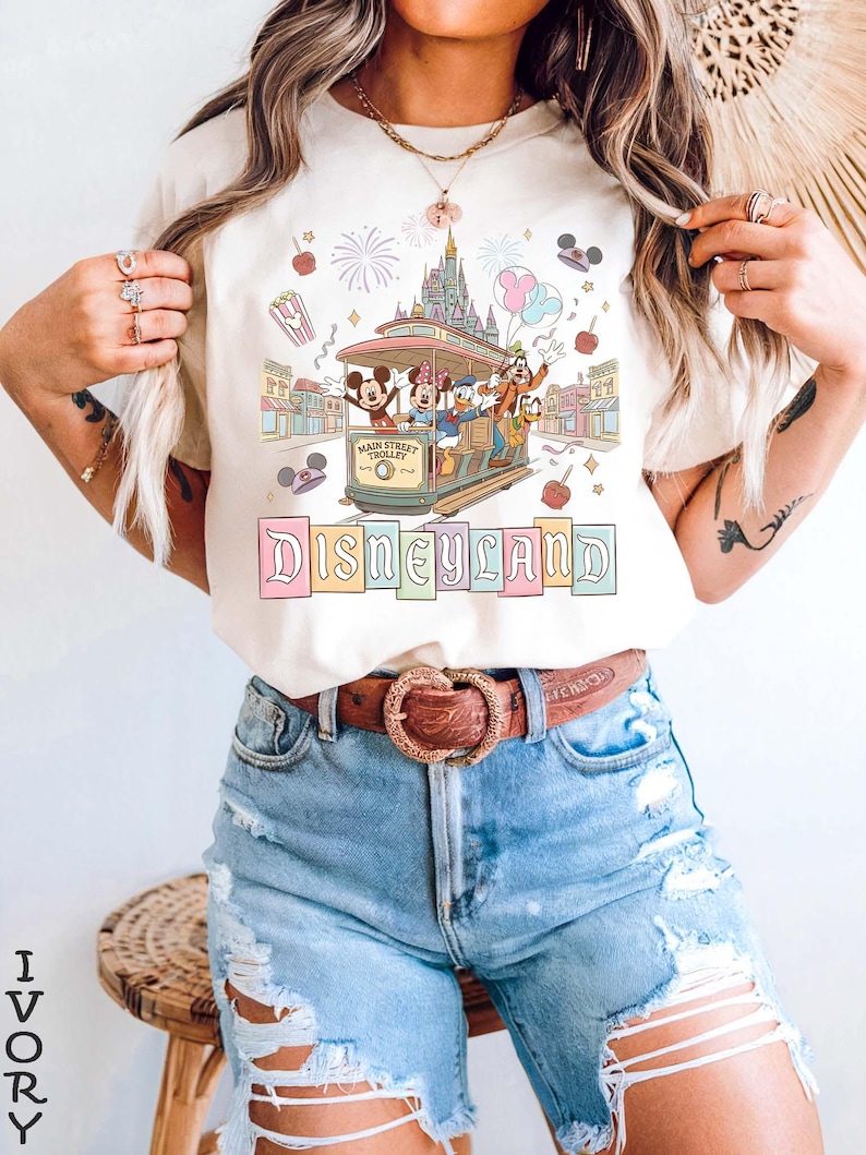 May include: Ivory-colored t-shirt featuring a Disneyland design. The graphic includes a trolley with Mickey, Minnie, Donald, Goofy, and Pluto, with the Disneyland castle in the background. The word "Disneyland" is written in pastel colors.