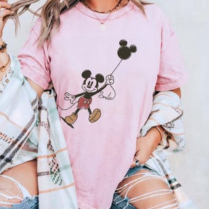 May include: A pale pink t-shirt with a Mickey Mouse graphic holding a black balloon. The word "Smile" is written below the balloon. The shirt is part of a clothing collection.
