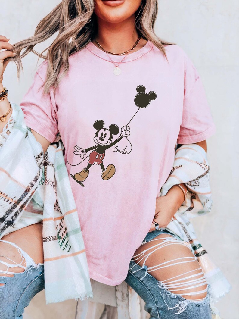May include: A pale pink t-shirt with a Mickey Mouse graphic holding a black balloon. The word "Smile" is written below the balloon. The shirt is part of a clothing collection.