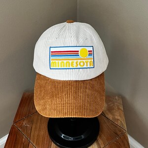 Op de afbeelding: Een witte corduroy baseballpet met een bruine corduroy klep. De voorkant van de pet heeft een geborduurd patch met een gele zon, blauwe en rode strepen en de tekst "MINNESOTA".