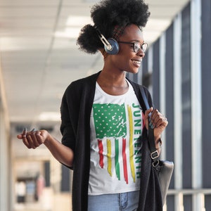 May include: A woman wearing a white t-shirt with a distressed American flag design and the text "Juneteenth" printed on the side. She is wearing a black cardigan, blue jeans, and has headphones on.