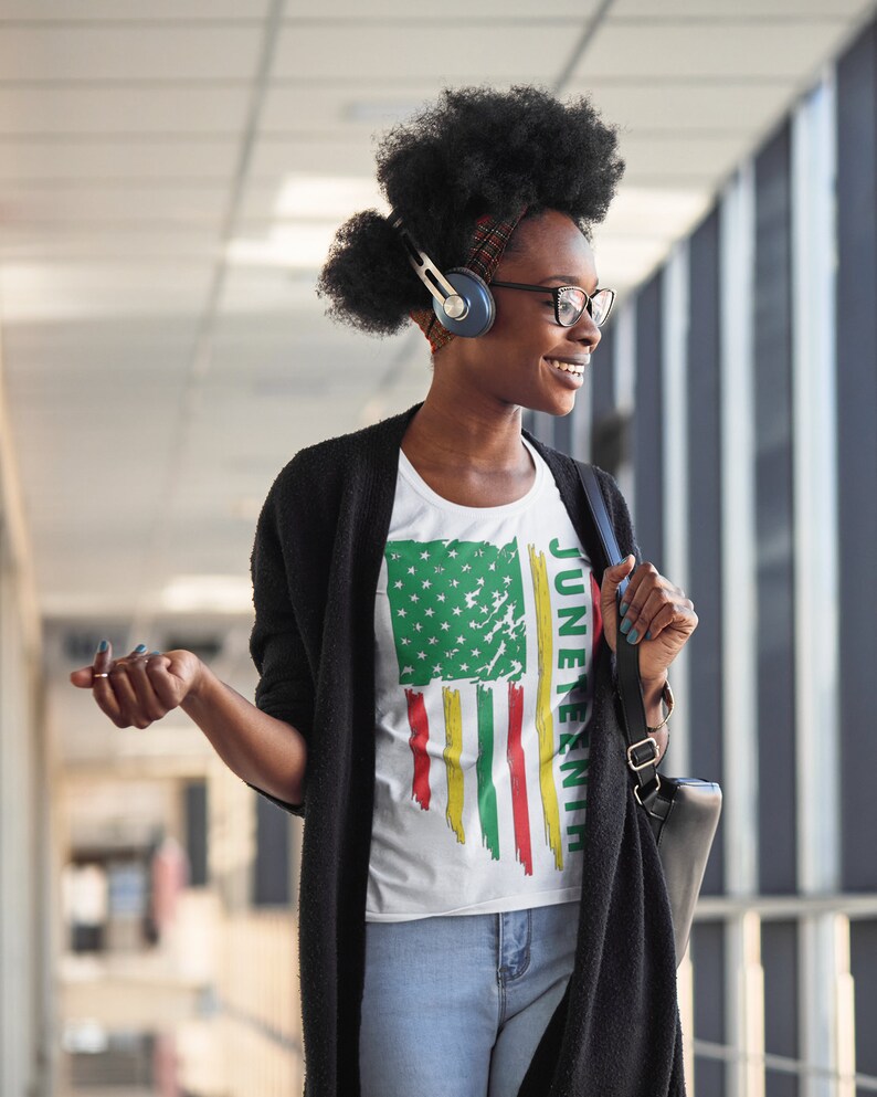 May include: A woman wearing a white t-shirt with a distressed American flag design and the text "Juneteenth" printed on the side. She is wearing a black cardigan, blue jeans, and has headphones on.