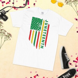 May include: White t-shirt featuring a distressed American flag design in green, red, and yellow. The word "Juneteenth" is written vertically on the right side of the flag.