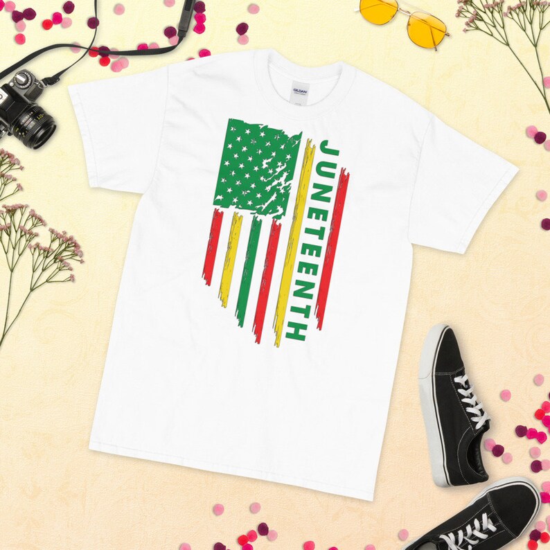 May include: White t-shirt featuring a distressed American flag design in green, red, and yellow. The word "Juneteenth" is written vertically on the right side of the flag.