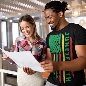 May include: A man and a woman are looking at a document. The man is wearing a black t-shirt with a green, yellow and red American flag design and the text "Juneteenth".