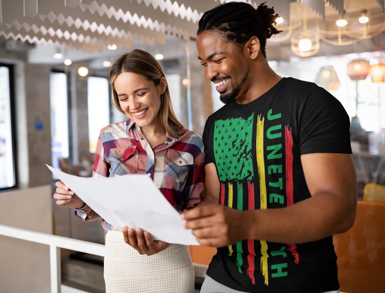 May include: A man and a woman are looking at a document. The man is wearing a black t-shirt with a green, yellow and red American flag design and the text "Juneteenth".