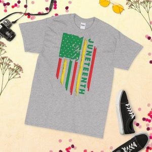 May include: A grey t-shirt featuring a distressed American flag design in green, yellow and red. The word "Juneteenth" is written vertically on the right side of the flag.
