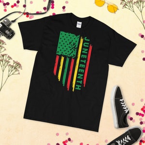 May include: Black t-shirt featuring a distressed American flag design in green, red and yellow. The text "Juneteenth" is written vertically on the right side of the flag.