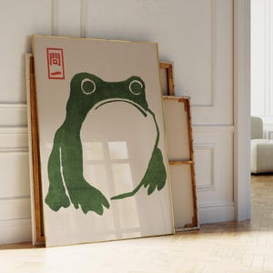 Puede incluir: Una impresión artística enmarcada con una ilustración de rana verde estilizada. La rana tiene ojos grandes y una boca ancha, sobre un fondo beige. La impresión está en un marco dorado, con un carácter japonés rojo en la esquina superior izquierda.