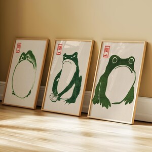 May include: Three framed art prints featuring green frog illustrations in a minimalist style. Each print has a wooden frame and a white background. The frog designs vary, with one holding a cup.