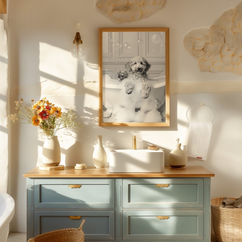 May include: A light blue bathroom vanity with two drawers and a white porcelain sink. A framed black and white photo of a dog in a bathtub with bubbles hangs above the sink. The vanity is made of wood and has gold hardware.