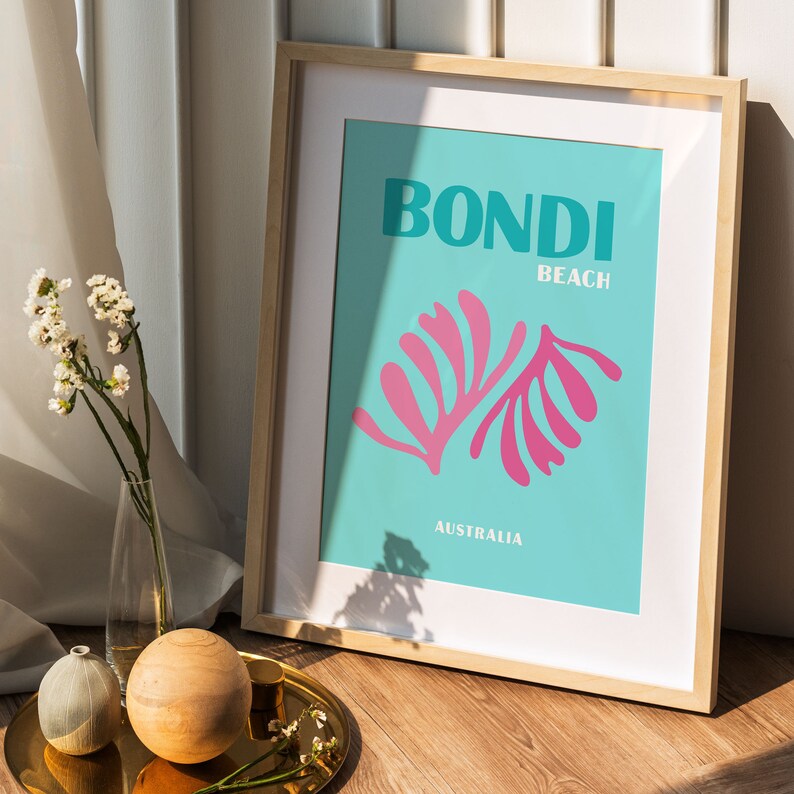May include: A framed print with a turquoise background and white text that reads "BONDI BEACH AUSTRALIA". The print features a pink stylized illustration of two fern-like plants.