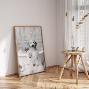May include: A black and white photograph of a dog in a bathtub full of bubbles. The dog is sitting in the tub with its paws on the edge. The photo is framed in a wooden frame.