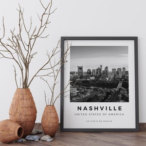 以下が含まれることがあります： 「NASHVILLE, UNITED STATES OF AMERICA, 36.1718&deg;N 86.7850&deg;W」というテキストが画像の下に印刷された、ナッシュビルのスカイラインの白黒写真。写真は黒いフレームで額装され、白い壁に飾られており、乾燥した枝が入った木製の壺が3つある。