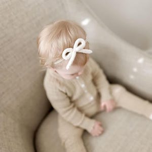 Op de afbeelding: Een witte gebreide strik hoofdband op het hoofd van een baby. De baby draagt een beige outfit en zit in een beige stoel. De hoofdband is het middelpunt, met de strik gecentreerd op het voorhoofd.