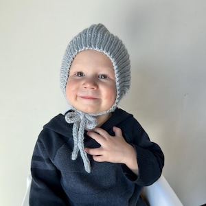 May include: A gray knitted baby bonnet with a pointed top and tie closure. The bonnet is made of a soft, ribbed knit material. The baby is wearing a dark blue hooded sweatshirt.