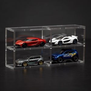 Puede incluir: Una vitrina de acrílico transparente con cuatro compartimentos, cada uno con un coche de juguete diferente. Los coches son un coche deportivo rojo, un coche deportivo blanco, un sedán gris y un SUV azul. La vitrina está sobre una superficie negra.