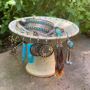 May include: A white ceramic dish with a scalloped edge holds a collection of turquoise and silver jewelry. The jewelry includes several bracelets, earrings, and a pendant. The dish is sitting on a stone surface.