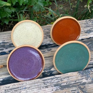 May include: Four ceramic coasters in different colors: green, purple, white with brown speckles, and orange. The coasters are round and have a slightly raised edge.