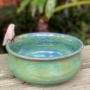 May include: A round ceramic bowl with a glossy, variegated glaze in shades of teal, turquoise, and lime green. A small, light-colored ceramic bird figurine with dark speckles is attached to the outer rim of the bowl, facing inward.