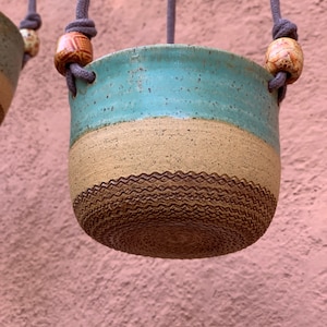 May include: A hanging planter made of brown speckled ceramic with a light blue and brown glaze. The planter is suspended by a gray rope with brown wooden beads.