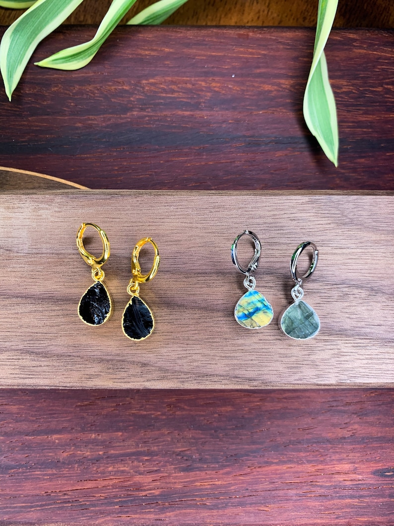 May include: Two pairs of drop earrings. The first pair features gold-tone hoops with black, teardrop-shaped stones. The second pair features silver-tone hoops with iridescent, teardrop-shaped stones displaying blue and yellow hues.