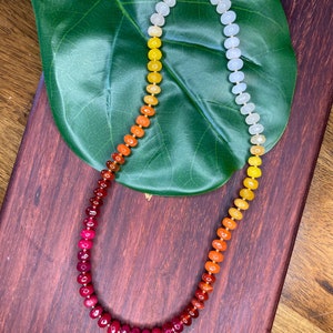 May include: A beaded necklace with a gradient of colors, transitioning from red to orange, yellow, and finally white. The necklace is displayed on a wooden surface with a large green leaf in the background. The necklace has a gold clasp.