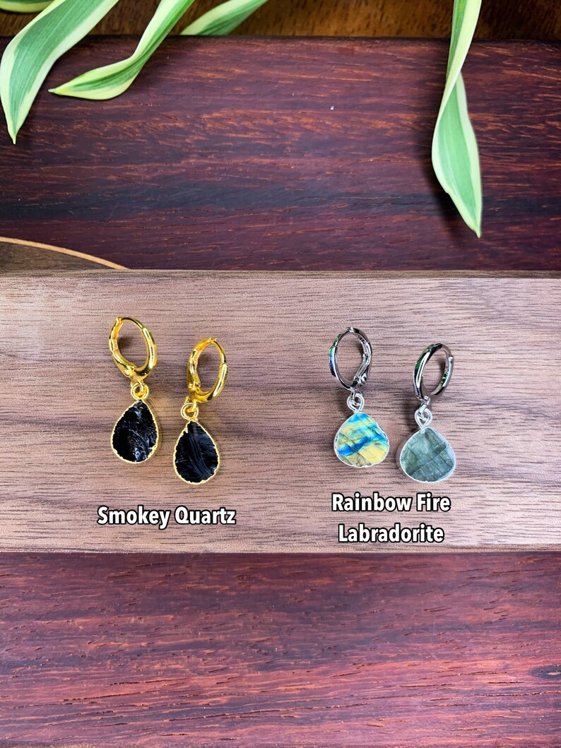 May include: Two pairs of hoop earrings with teardrop-shaped gemstone pendants. The left pair features black Smokey Quartz stones on gold-toned hoops, while the right pair features iridescent Rainbow Fire Labradorite stones on silver-toned hoops.