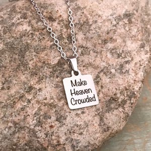 May include: A silver-colored necklace with a square pendant. The pendant has the words "Make Heaven Crowded" engraved in black. The necklace is displayed on a textured, light brown stone surface.