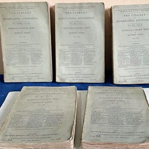 Könnte beinhalten: Eine Sammlung antiker Bücher mit dem Titel "The Library, Entertaining Knowledge, Distinguished Men of Modern Times". Die Bücher sind in einem verblichenen Grauton gebunden, mit schwarzem Text. Die Bücher sind auf einer blauen Oberfläche angeordnet.