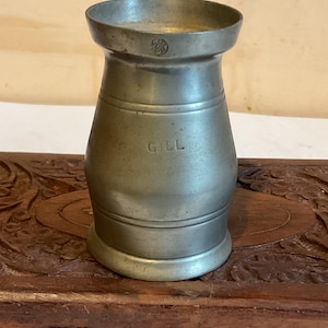 K&ouml;nnte beinhalten: Ein silberner Zinnmessbecher mit der Aufschrift "Gill" an der Seite. Der Becher steht auf einer Holzoberfl&auml;che.