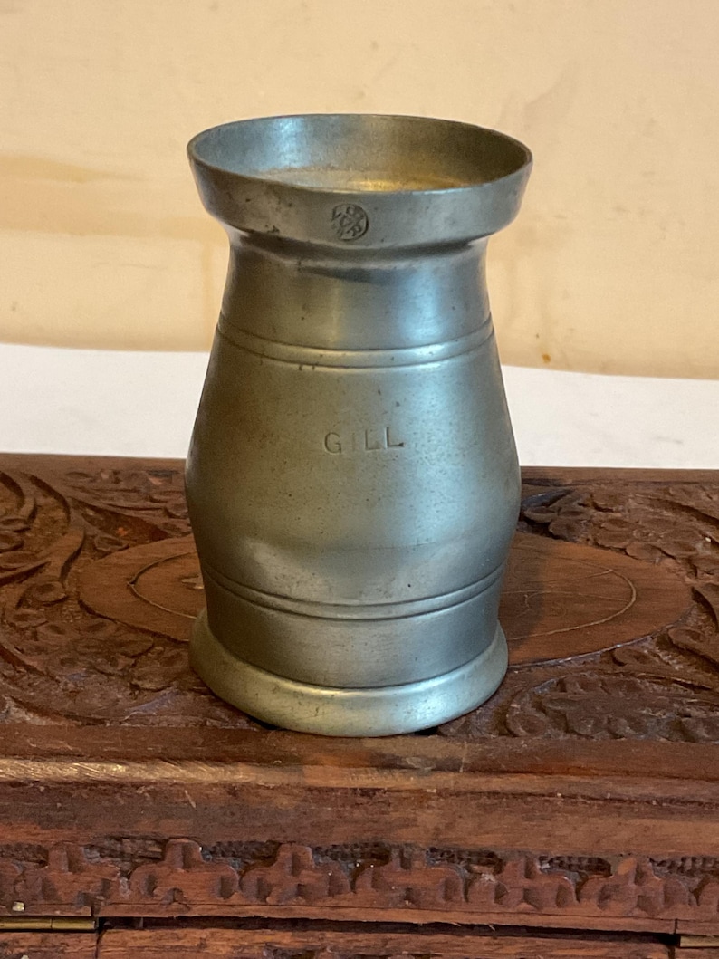 K&ouml;nnte beinhalten: Ein silberner Zinnmessbecher mit der Aufschrift "Gill" an der Seite. Der Becher steht auf einer Holzoberfl&auml;che.