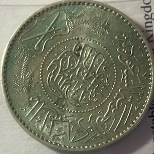 Könnte beinhalten: Eine silberne Münze mit aufwendiger arabischer Schrift und dekorativen Elementen. Die Münze zeigt ein zentrales Design, umgeben von einem Perlenrand und kunstvollen Mustern. Die Oberfläche der Münze weist Gebrauchsspuren auf.
