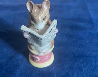 May include: A ceramic figurine of a mouse reading a newspaper. The mouse is brown and white, perched atop a spool of thread. The newspaper reads "Sailor" and "Cutter". The spool is pink and yellow.