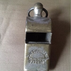 Puede incluir: Un silbato plateado con un anillo negro y una parte superior plateada. El silbato tiene el texto "The Thunderer Hudson's Patent" grabado en el lateral.