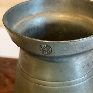 K&ouml;nnte beinhalten: Eine silberne Zinnkanne mit einer kreisf&ouml;rmigen Markierung an der Seite. Die Markierung ist ein Kreis mit einem Kreuz darin.