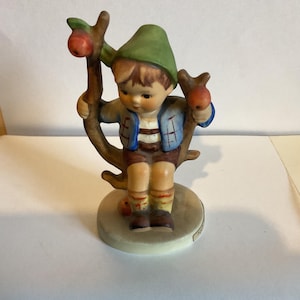 Op de afbeelding: Een porseleinen beeldje van een jongen met een groene hoed en een blauw shirt, zittend op een tak met appels. Het beeldje staat op een witte voet met de handtekening van de kunstenaar.