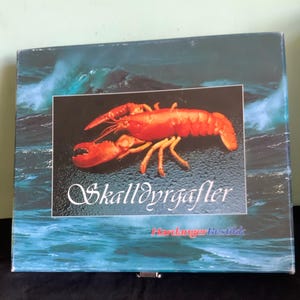 May include: A square box with a dark blue background depicting ocean waves. A rectangular image in the center features a bright red lobster on a black background. The text "Skalldyrgafler" is written in white script.