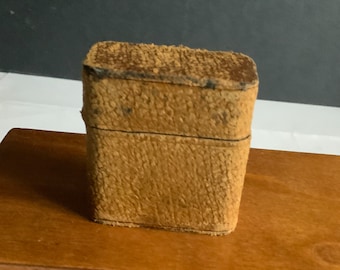 May include: A small, rectangular box made of brown leather with a textured surface. The box has a hinged lid and a small latch.