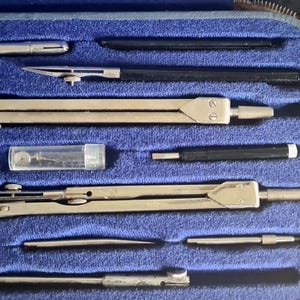 Könnte beinhalten: Ein Vintage-Zeichensatz in einem blau mit Samt gefütterten Etui. Das Set enthält mehrere Metallzirkel, Teiler und andere Zeichenwerkzeuge. Die Werkzeuge sind silberfarben und ordentlich im Etui angeordnet.