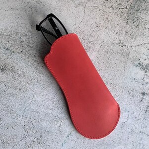 Puede incluir: Un estuche para gafas de cuero rojo con un par de gafas negras en su interior.