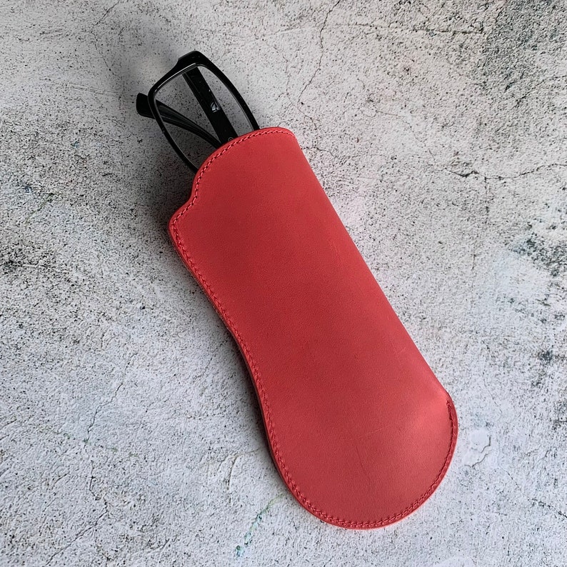 Puede incluir: Un estuche para gafas de cuero rojo con un par de gafas negras en su interior.