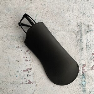 Puede incluir: Un estuche para gafas de cuero negro con un par de gafas negras en su interior.